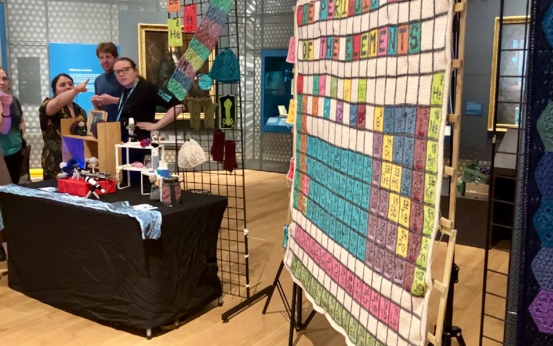 Image of a stall at the "Lates" event. On the left-hand side of the image, a group of five people can be seen near a black stall that is decorated with science crafts. One member of the group is pointing towards the right-hand side of the image where there is a wooly periodic table hanging up.