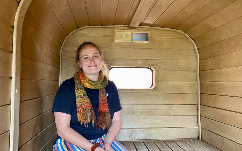 Saorla Wright, a 27-year-old grief sauna host, sits in a wooden sauna with a smile on her face. She is wearing a navy top, striped pants, and an autumnal-coloured scarf