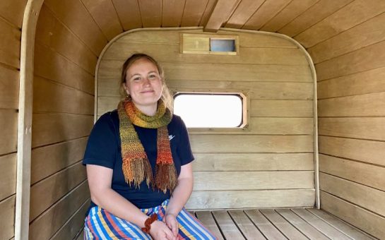 Saorla Wright, a 27-year-old grief sauna host, sits in a wooden sauna with a smile on her face. She is wearing a navy top, striped pants, and an autumnal-coloured scarf