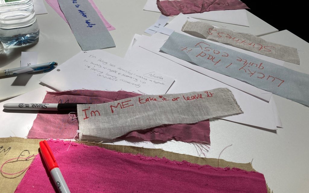 Scraps of fabric on a table, with words written on it in marker pen. Scrap in the centre reads "I’m me, take it or leave it."