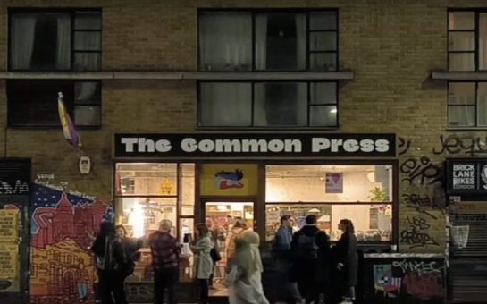 People stand at night time outside a brightly-lit bookshop with the words 'The Common Press' written in white text on a black background above the windows.