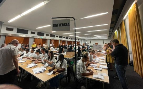 A picture from an election count of people counting votes. A sign saying Battersea is clearly visible.