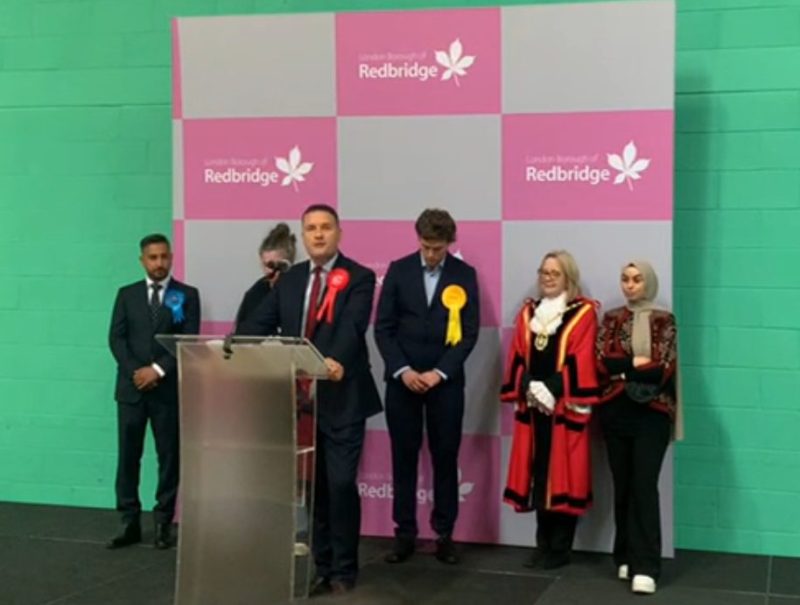 Politician Wes Streeting stands at a podium on a black stage. He is flanked by other Ilford North candidates. There is a pink and grey checkered board with 'Redbridge' logos behind them.