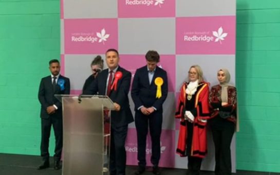 Politician Wes Streeting stands at a podium on a black stage. He is flanked by other Ilford North candidates. There is a pink and grey checkered board with 'Redbridge' logos behind them.