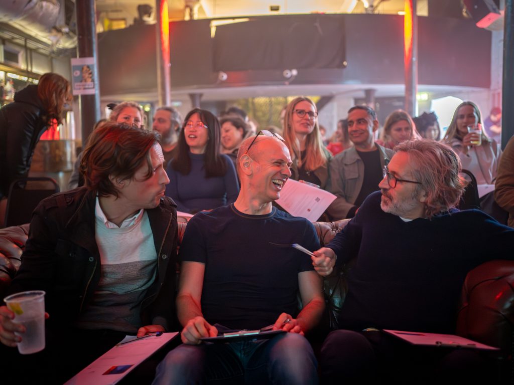 Three men laugh sitting on a brown leather sofa with a crowd of people sitting in rows behind them.