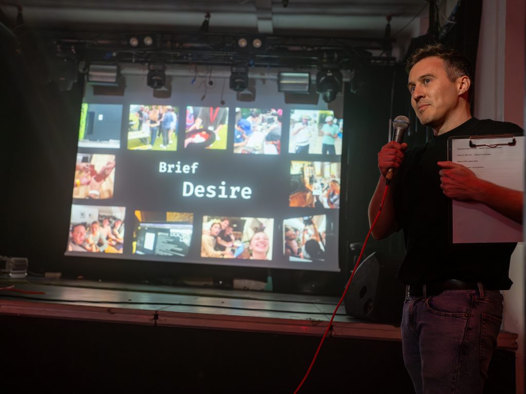 A man with a mic stands in front of a screen that says "Brief: Desire".