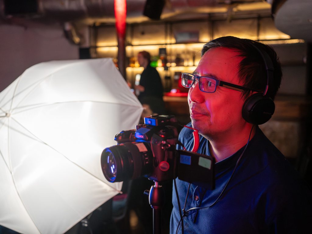 A man in glasses and headphones stands in front of a camera while interviewing somebody.