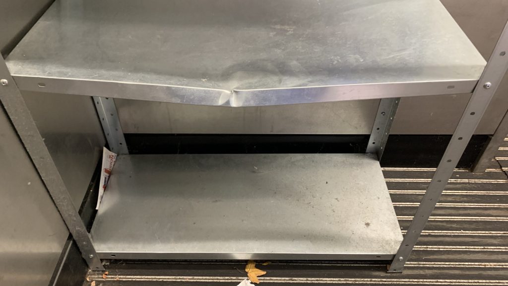 The image shows a metal bookshelf at Haggerston station's community book area. with a visible inward dent in the middle of the third shelf.