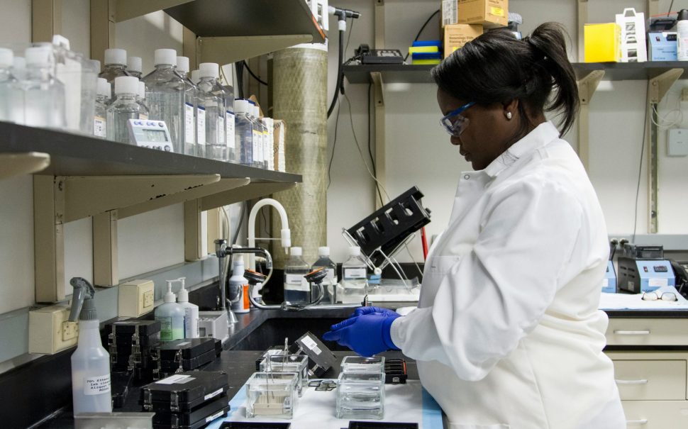 An image of a woman in a white lab technician coat standing over a sink in the lab.