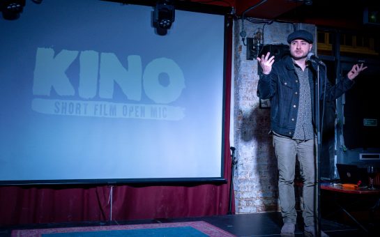 A man stands on a stage in a dark room addressing a film audience with the words: 'Kino Short Film Open Mic' in the background.